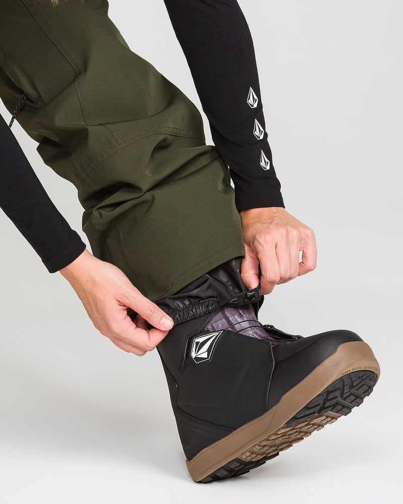 A person in Volcom Women’s Pollock Baggy Pants and a black long-sleeve top adjusts a black snow boot with a brown sole and geometric logo, preparing for outdoor winter activity.