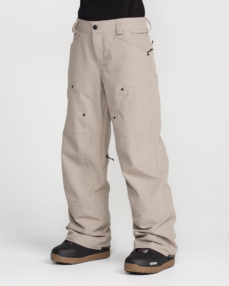 A person wearing Volcom Women’s Pollock Baggy Pants in light beige, featuring multiple pockets and visible seams, paired with black snow boots. The loose-fitting pants are shown from the waist down against a plain white background.
