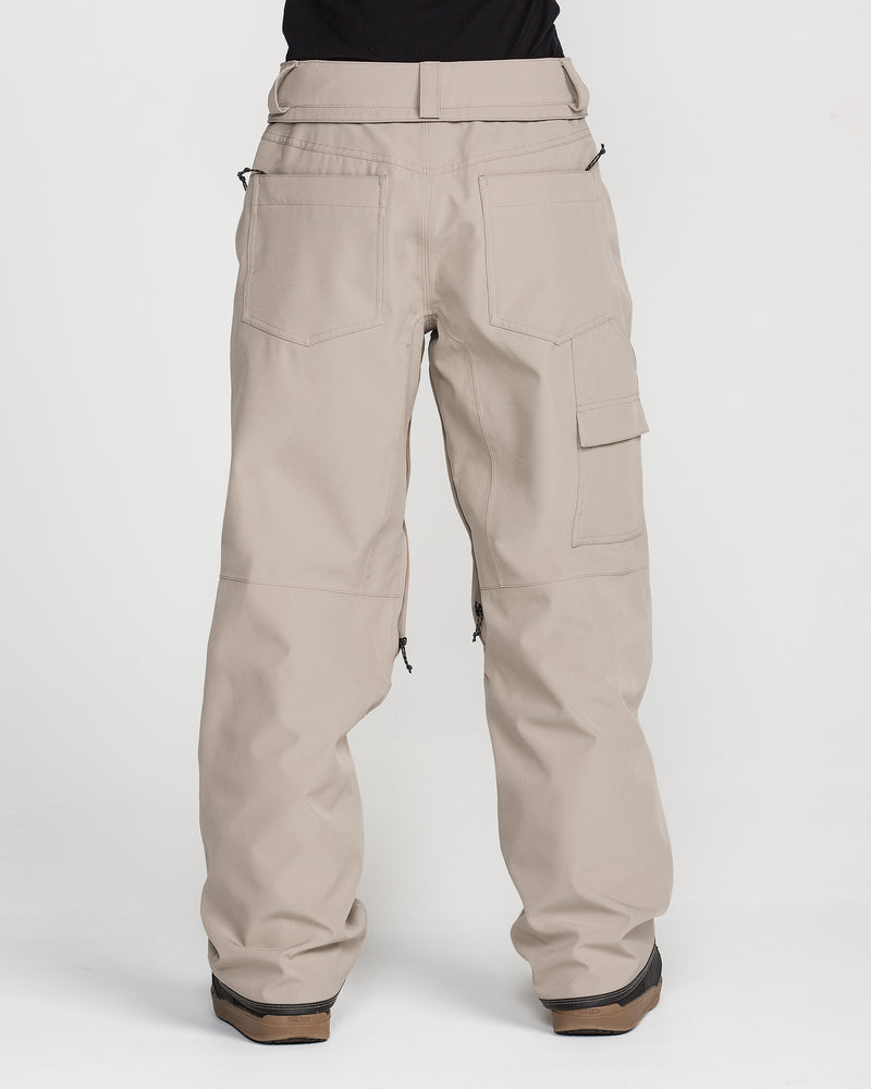A person is shown from the back, standing on a white background, wearing Volcom Women’s Pollock Baggy Pants in beige. The pants feature multiple pockets, and brown-soled shoes are visible.