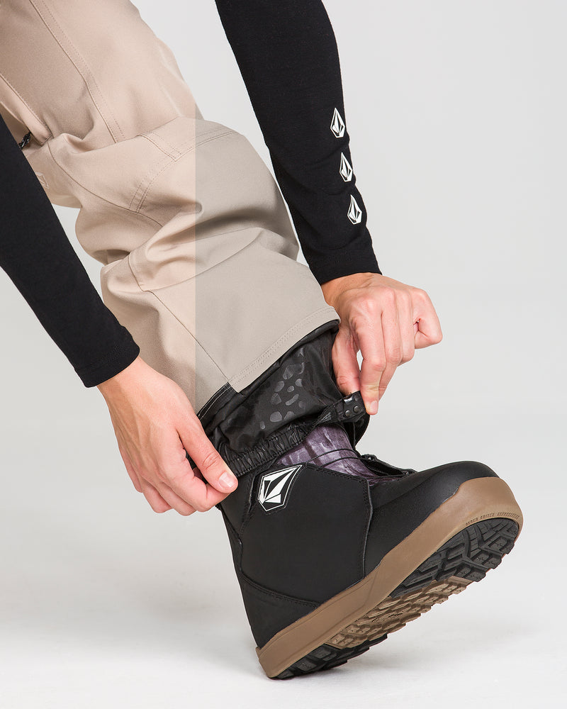 A person tucks beige Volcom Women’s Pollock Baggy Pants with a black gaiter into a black snow boot featuring a brown sole and a white logo. The person’s hand is adjusting the gaiter, and their sleeve displays three white logos.