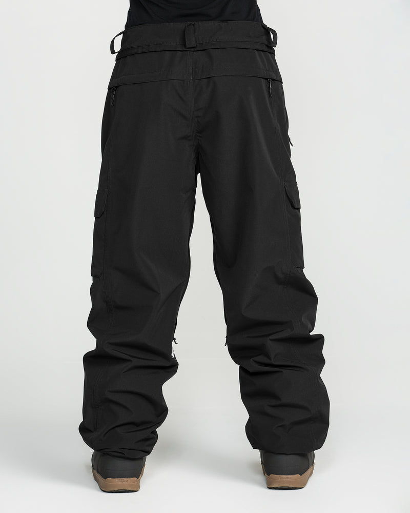 A person wearing loose-fitting black Volcom Unisex Liftline 20K Pants, made from recycled ski pant fabric, paired with brown boots and standing against a plain white background.