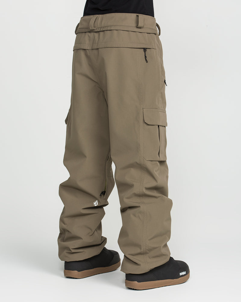 A person wears Volcom Unisex Liftline 20K Pants—loose khaki cargo pants made from recycled fabric with zipper pockets—paired with black slip-on shoes, standing against a white background. The image shows the lower half of their body from the back.