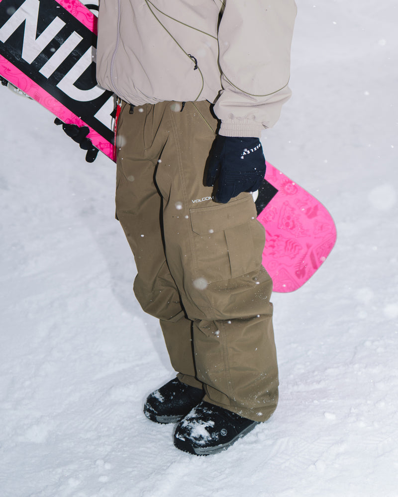 A person in Volcom Unisex Liftline 20K Pants standing in the snow and holding a pink snowboard.