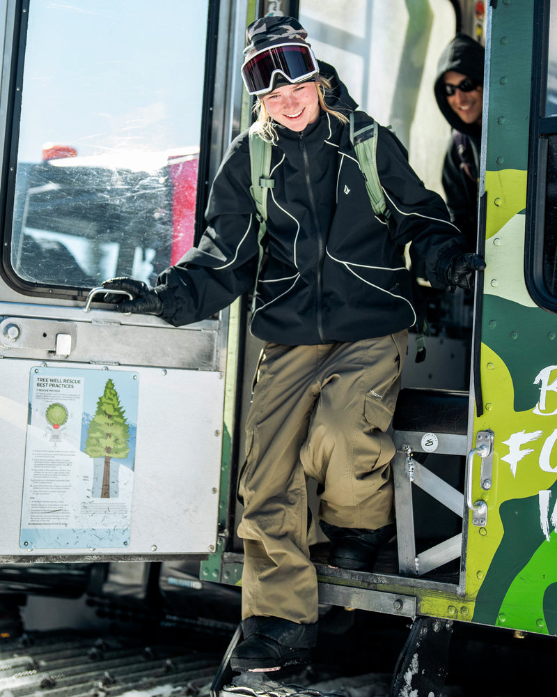 A smiling person wearing snow goggles and a black jacket steps out of a snowcat in olive green Volcom Unisex Liftline 20K pants.