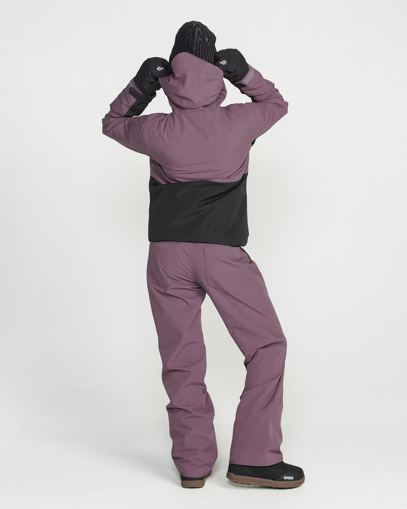 A person stands facing away against a plain white background, wearing a purple and black hooded jacket, black gloves, black shoes, and Volcom Women’s Stoned Pants.