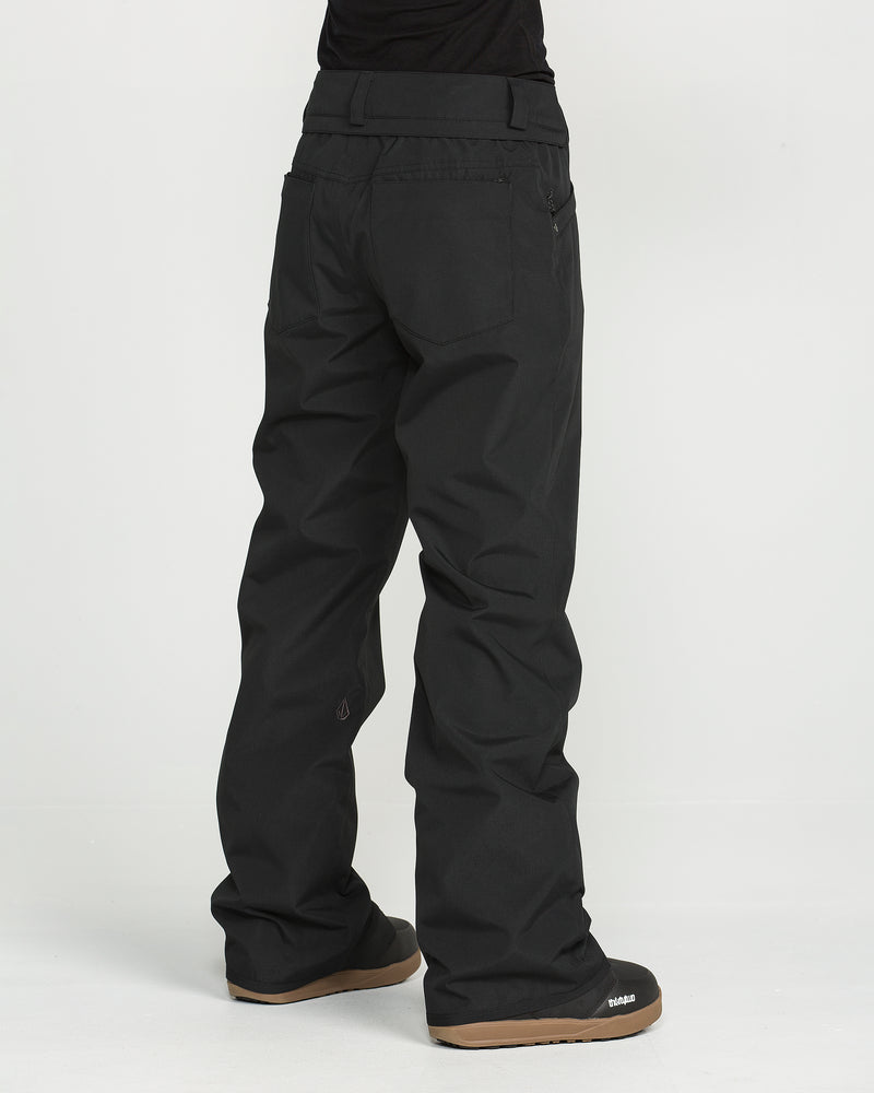 A person is seen from behind against a plain white background, wearing Volcom Women’s Stoned Pants—loose-fitting black snow pants featuring V-Science 2-Layer technology—paired with black boots.