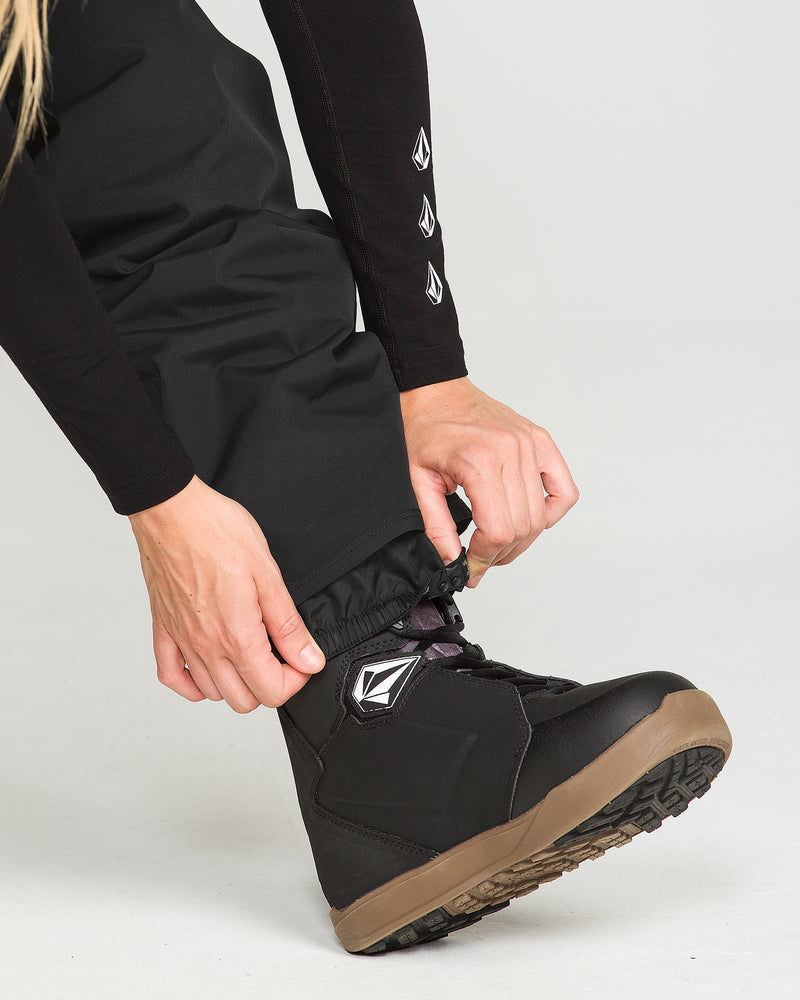 Wearing Volcom Women’s Stoned Pants in black and a matching black long-sleeve top, a person adjusts the drawstring at the pant leg, which is styled over black snow boots with brown soles.