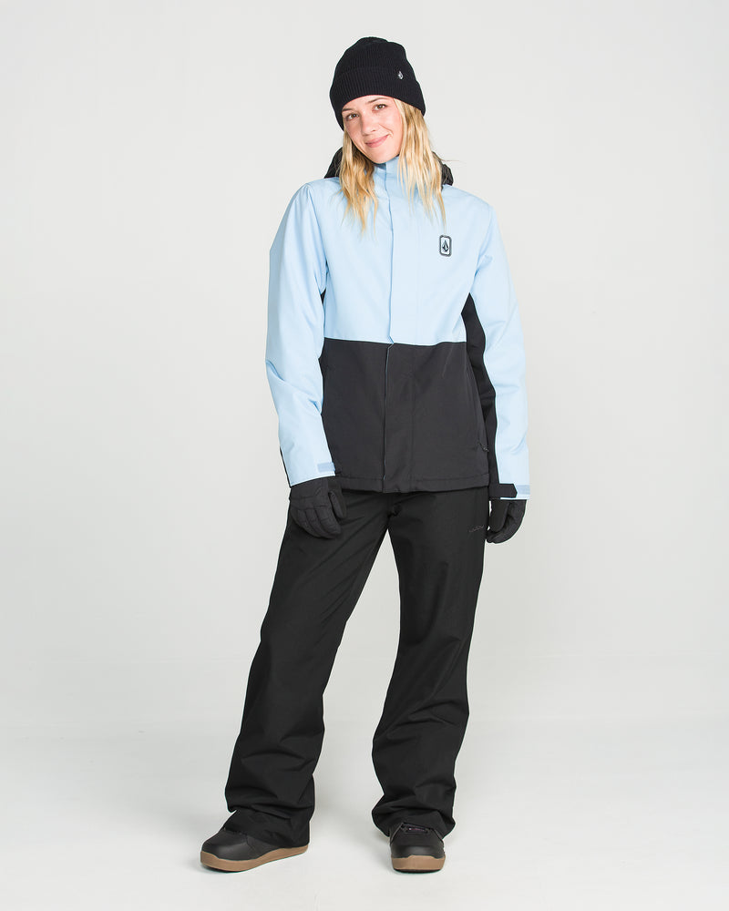 A person wearing black Volcom Women’s Stoned Pants, a light blue and black winter jacket, gloves, boots, and a black beanie stands in a studio with a white background, looking at the camera and smiling slightly.