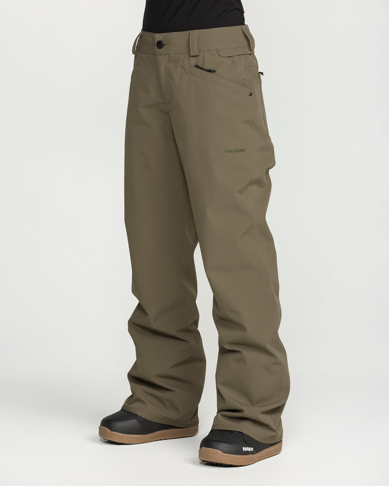A person is shown from the waist down, standing against a plain white background, wearing Volcom Women’s Stoned Pants in olive green. The waterproof snow pants feature a relaxed fit and display a small logo on the left thigh. Black snow boots complete the look.