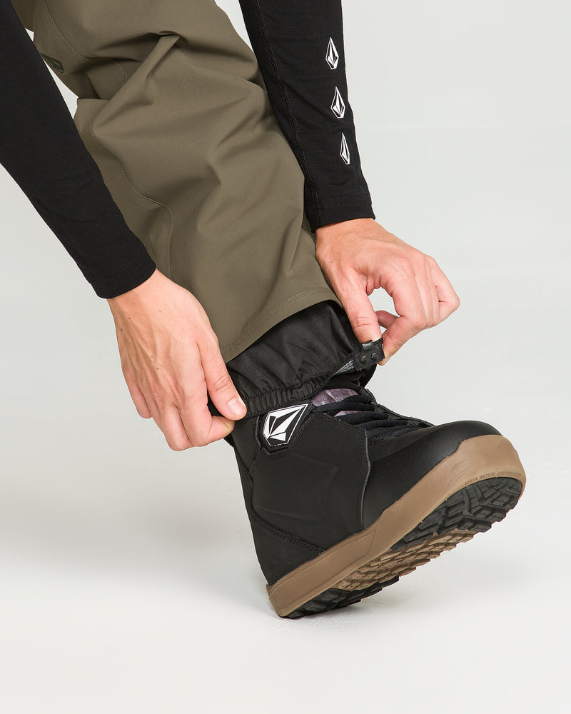 A person wearing Volcom Women’s Stoned Pants in olive and a black long sleeve shirt tightens the elastic cuff over a black snow boot with a brown sole.