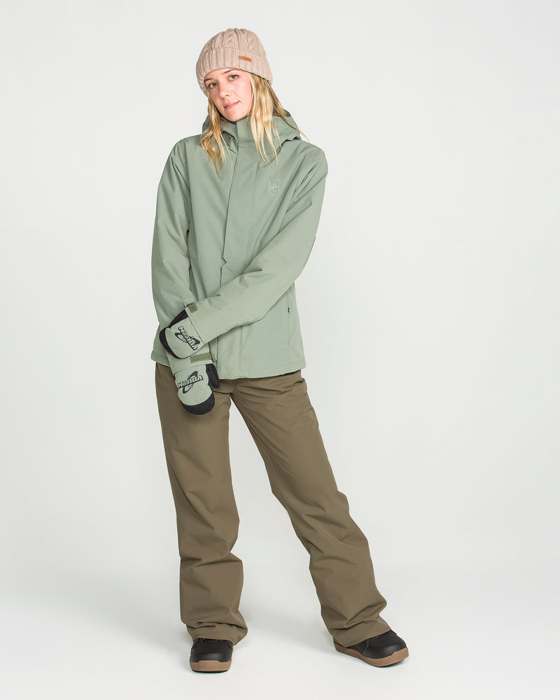 A person wearing a green hooded jacket, Volcom Women’s Stoned Pants, black boots, gray mittens, and a beige knit beanie stands against a plain white background, looking relaxed with their hands together.