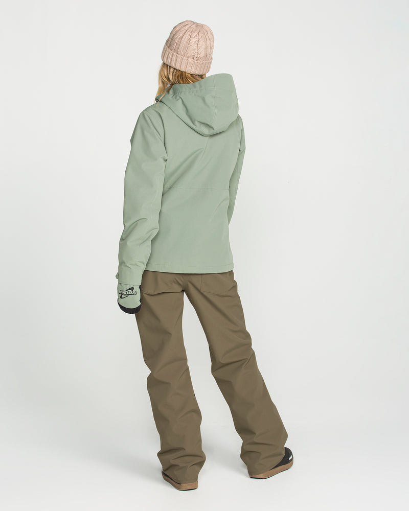 A person is seen from behind against a plain white background, wearing Volcom Women’s Stoned Pants in brown, a light green hooded jacket, black gloves, and a pink knit beanie.