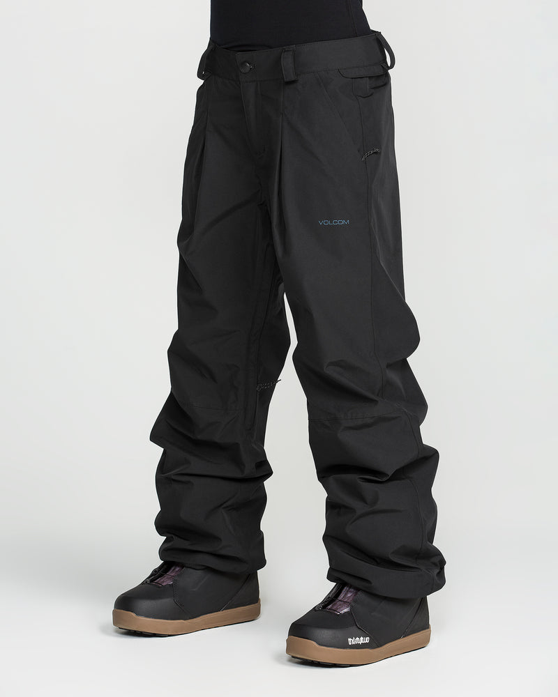A person wearing Volcom Unisex Yourfav Baggy Gore Pants in black stands against a plain white background, paired with black snow boots featuring brown soles. Only the lower body is visible.