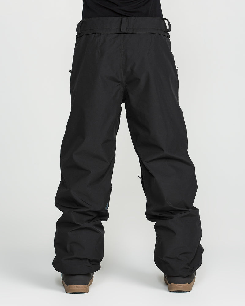 A person is wearing Volcom Unisex Yourfav Baggy Gore Pants in black, featuring an adjustable waist, along with black snow boots. They are standing against a plain white background, and their upper body is not visible in the image.