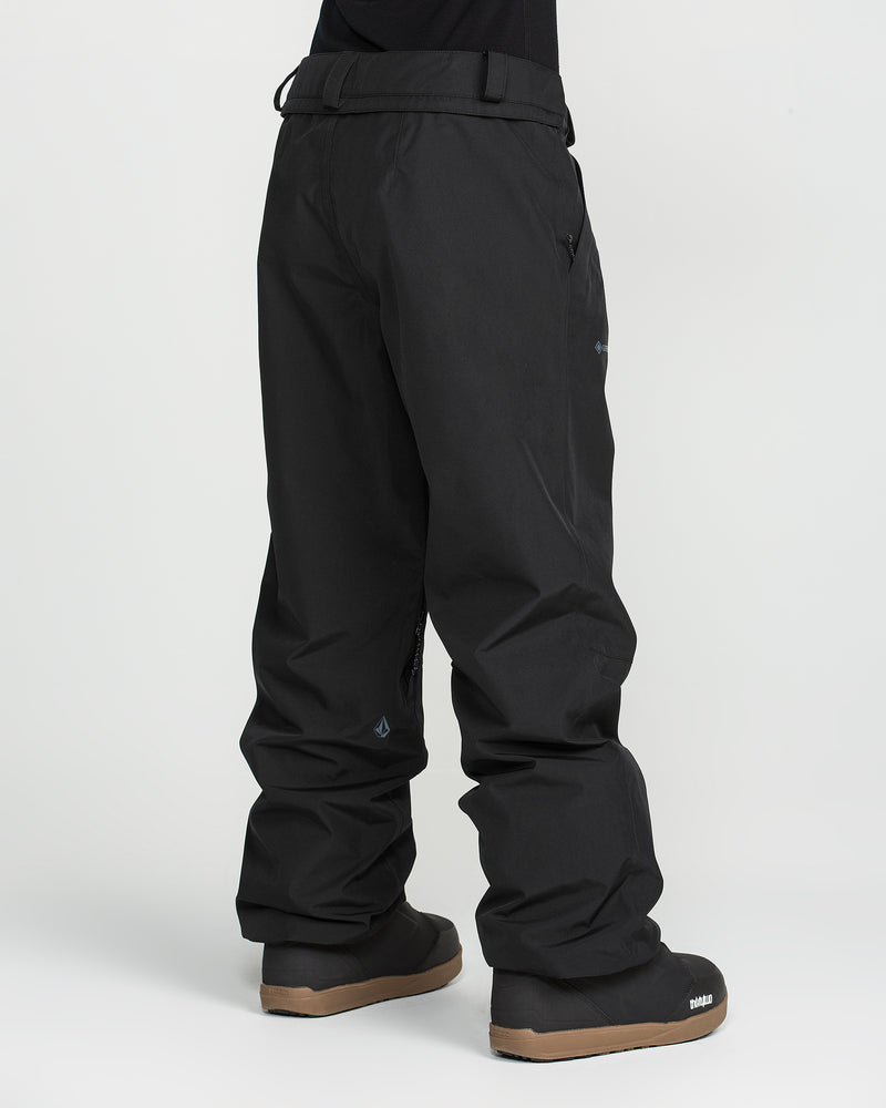 A person wearing Volcom Unisex Yourfav Baggy Gore Pants in black, featuring an adjustable waist, paired with black boots with brown soles, stands against a plain white background.