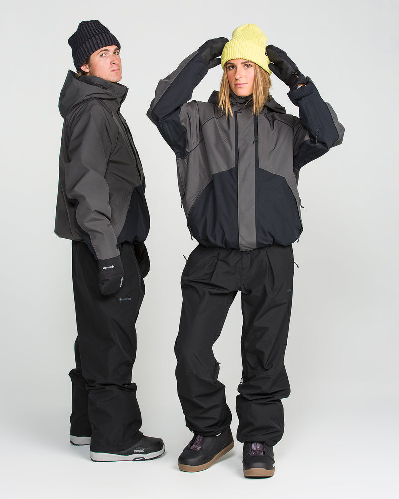 Two people dressed in black and gray winter outfits stand together against a white background. One adjusts a yellow beanie while the other wears a black beanie. Both are wearing gloves, jackets, winter boots, and the Volcom Unisex Yourfav Baggy Gore Pants—baggy pants with an adjustable waist made from recycled GORE-TEX fabric for extra comfort and sustainability.