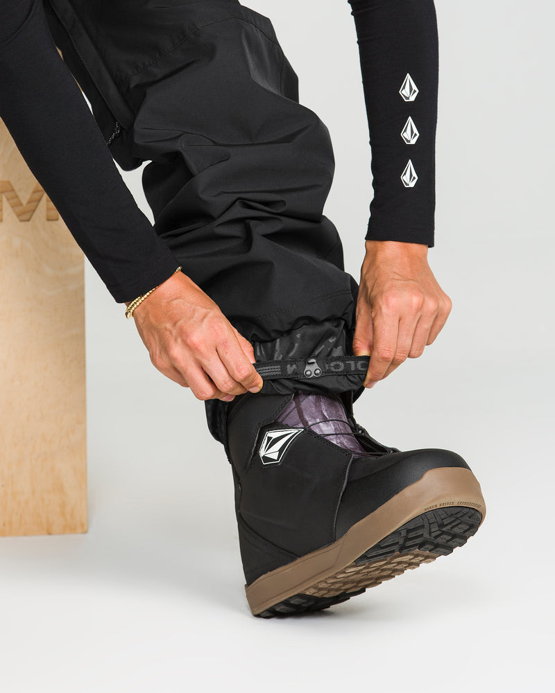 A person in black Volcom Unisex Yourfav Baggy Gore Pants and boots adjusts the cuff of the pants over the boot. These pants, made from recycled fabric, feature an adjustable waist and display the Volcom logo. The boots are black with a brown sole, set against a plain background.