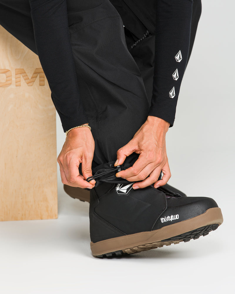 A person wearing Volcom Unisex Yourfav Baggy Gore Pants in black, featuring an adjustable waist, and a long-sleeve shirt adjusts the cuff over a black snow boot with a brown sole. The individual is seated on a wooden bench, and the boot displays the “thirtytwo” branding and logo.