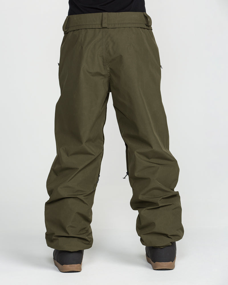 A person is shown from the waist down against a plain white background, wearing loose-fitting olive green Volcom Unisex Yourfav Baggy Gore Pants with an adjustable waist and black shoes.