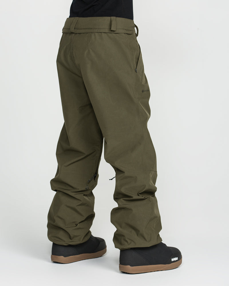 A person wearing Volcom Unisex Yourfav Baggy Gore Pants—olive green, loose-fitting outdoor pants with an adjustable waist and zippered pockets, slightly gathered at the ankles and made from durable recycled fabric—paired with black boots, stands against a plain white background.