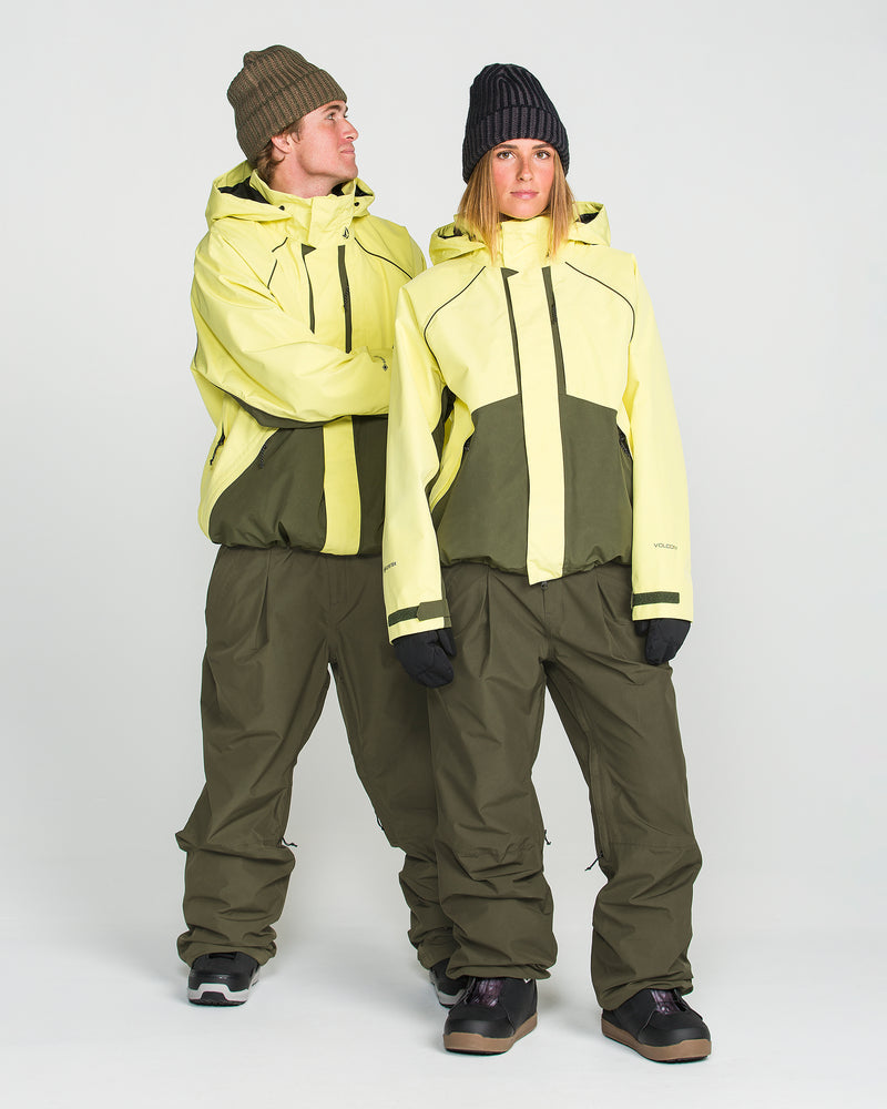 Two people wearing matching yellow and olive green winter jackets, black gloves, and beanies stand side by side and look at each other against a plain white background. They are both wearing the Volcom Unisex Yourfav Baggy Gore Pants in olive green with an adjustable waist.