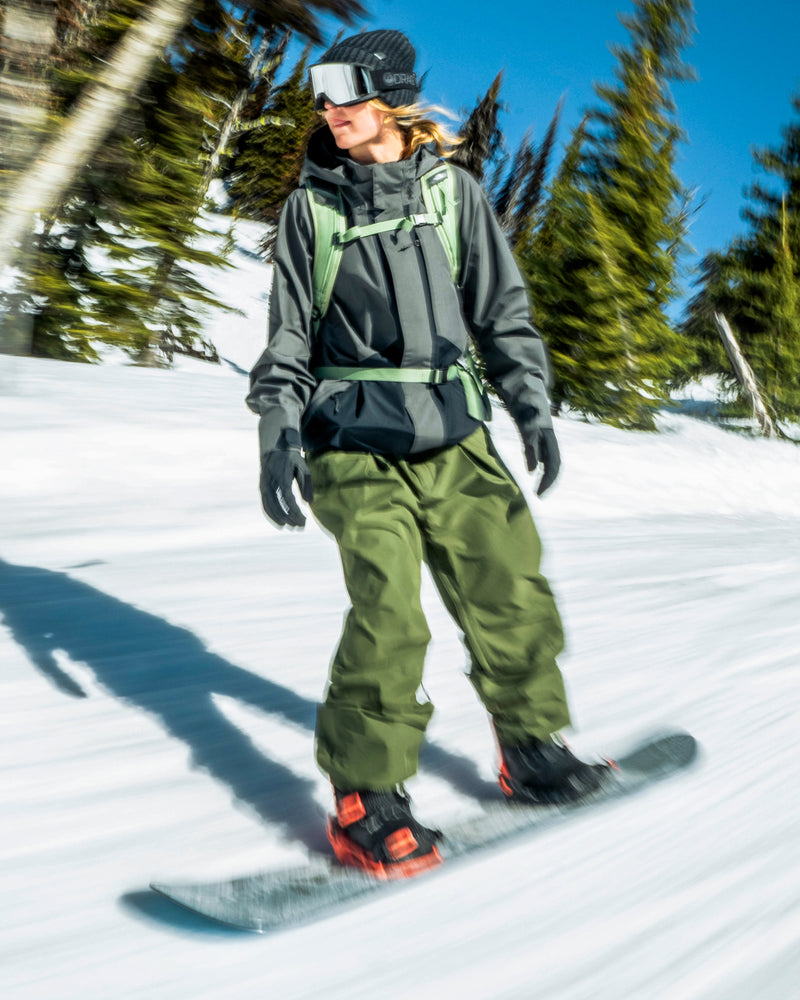 A snowboarder wearing a dark jacket, olive green baggy Volcom Unisex Yourfav Baggy Gore Pants, and a backpack rides down a snowy slope.