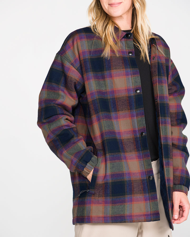 A woman models the Volcom Women’s Insulated Riding Flannel Jacket, featuring a loose fit in blue, green, and red plaid with a soft taffeta lining, layered over a black shirt and beige pants. She stands against a plain white background with a slight smile.
