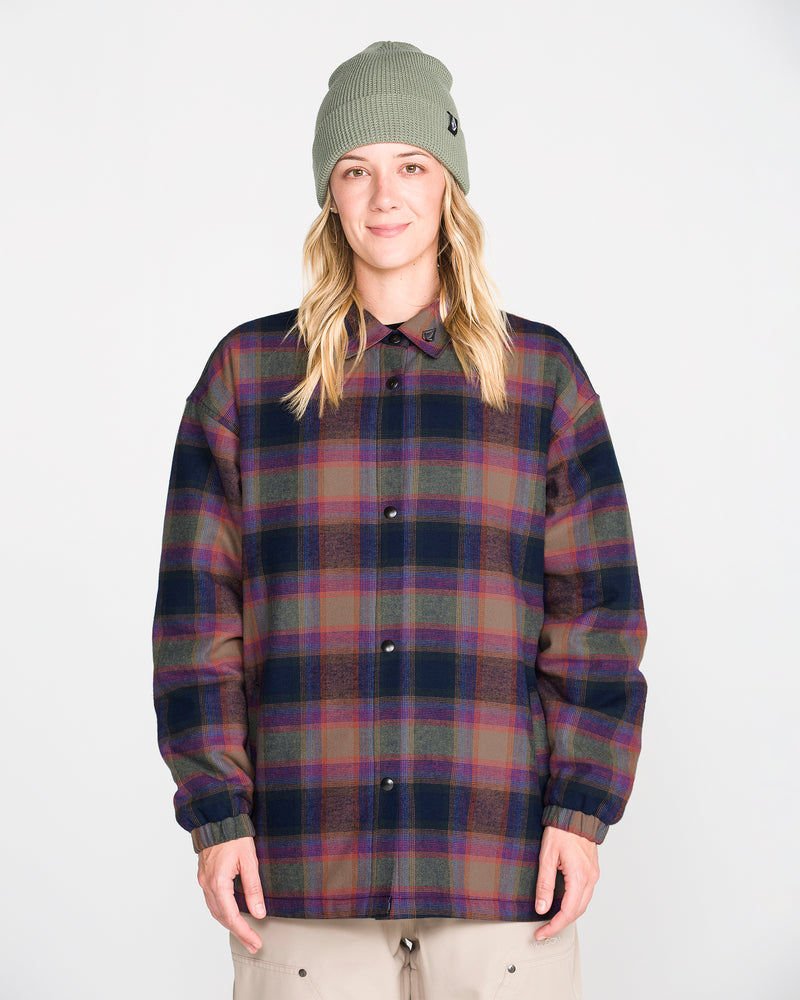 A person stands facing forward against a plain white background, hands relaxed by their sides, wearing the Volcom Women’s Insulated Riding Flannel Jacket—a plaid flannel jacket with taffeta lining—and a green knit beanie.