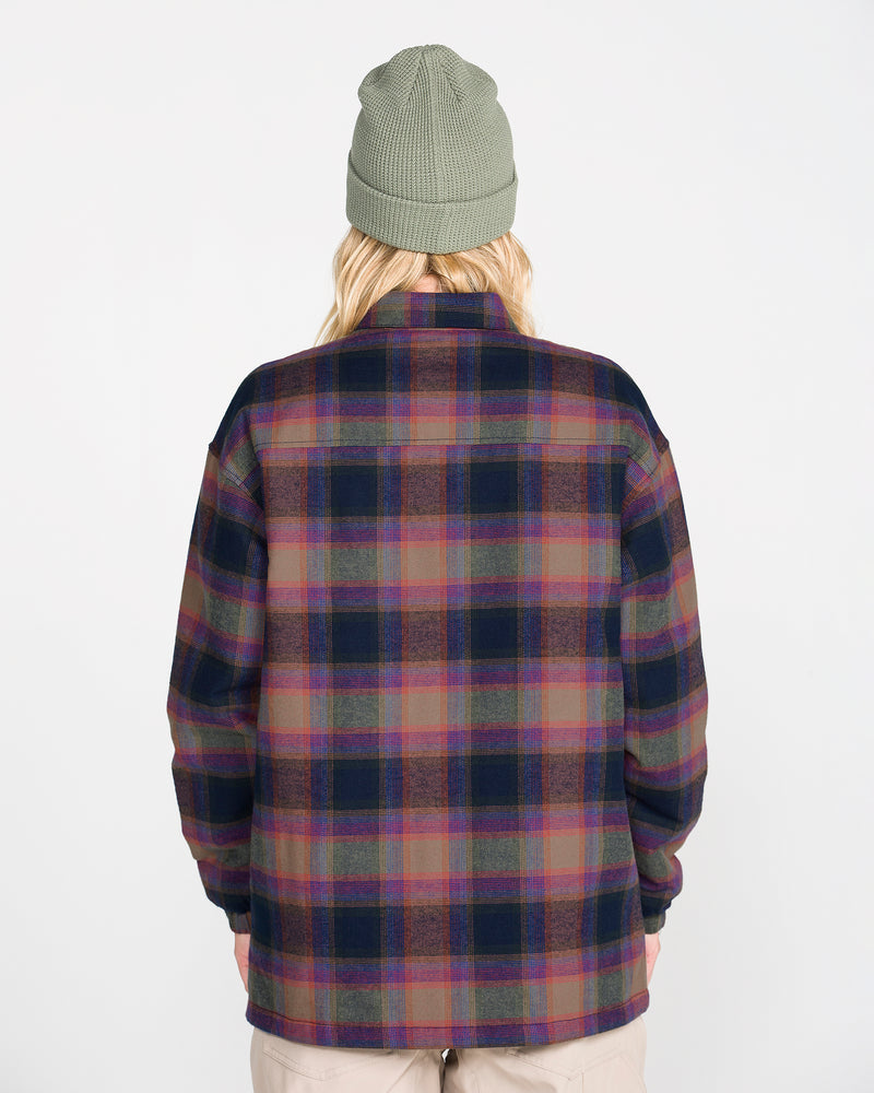 A person is shown from behind, wearing a green knit beanie and the Volcom Women’s Insulated Riding Flannel Jacket in a plaid pattern with shades of purple, pink, and green. The jacket features a soft taffeta lining for added comfort. The background is plain white.