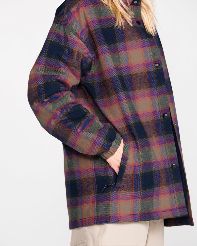 A person, with hand in pocket, models the Volcom Women’s Insulated Riding Flannel Jacket—a long, plaid outer layer in dark blue, green, and reddish-brown shades. The jacket is finished with a cozy taffeta lining. Only part of the person’s face and their light-colored pants are visible.