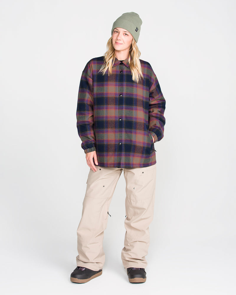 A person is seen standing facing forward against a plain white background, with hands in the pockets of a Volcom Women’s Insulated Riding Flannel Jacket featuring plaid flannel and taffeta lining, paired with a green beanie and beige pants.