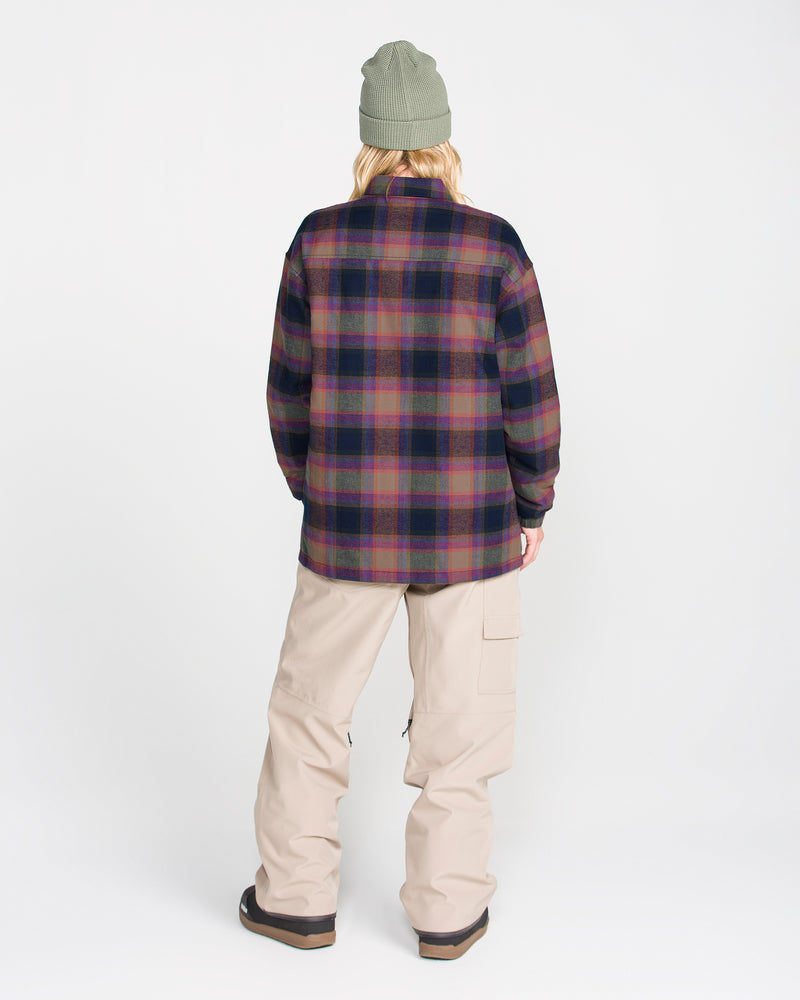 A person is seen from behind, standing against a plain white background, wearing the Volcom Women’s Insulated Riding Flannel Jacket in dark tones with taffeta lining, paired with a green beanie and loose beige pants featuring pockets.