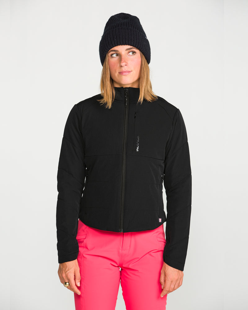 A woman wearing the Volcom Women’s Evergreen Full Zip Jacket in black, paired with a black beanie and bright pink pants, stands against a plain white background, gazing slightly to the side.