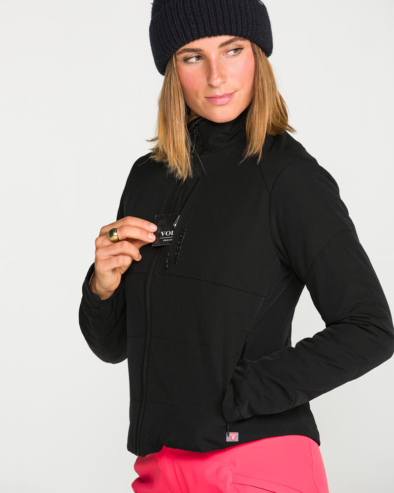 A woman wearing the Volcom Women’s Evergreen Full Zip Jacket and pink pants, with a black knit hat, holds a small black card near her chest pocket while looking to the side.