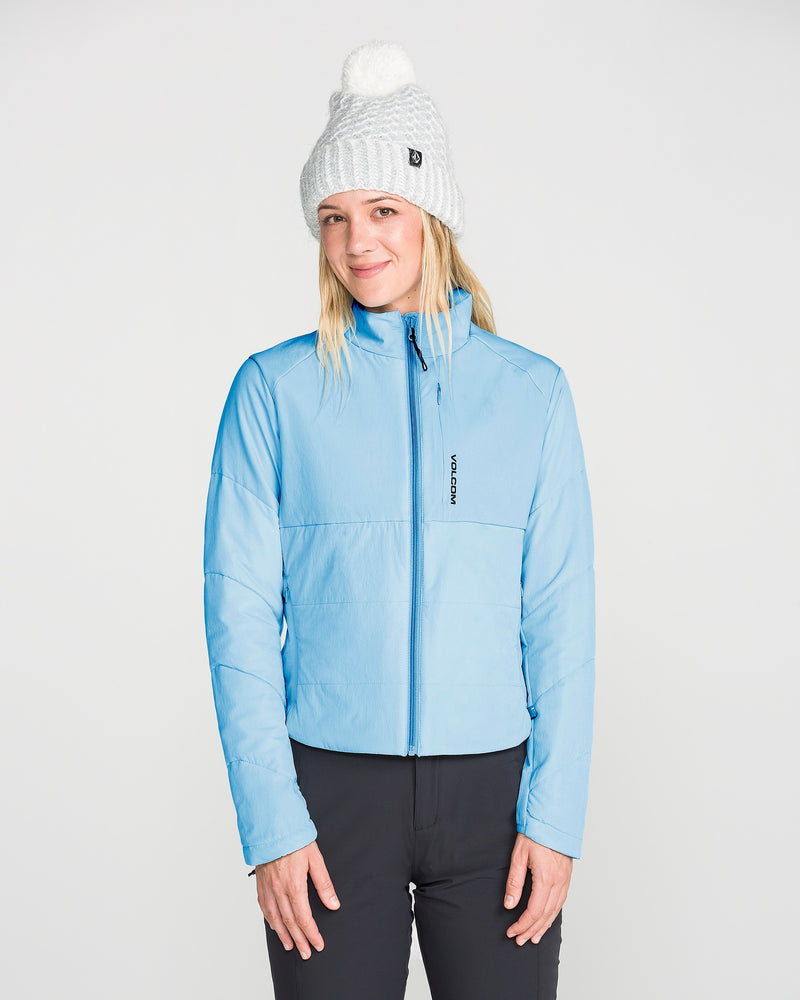A woman stands against a plain light background, smiling slightly, dressed in the Volcom Women’s Evergreen Full Zip Jacket in light blue—showcasing subtle quilted details and recycled insulation—paired with black pants and a white knit beanie topped with a pom-pom.
