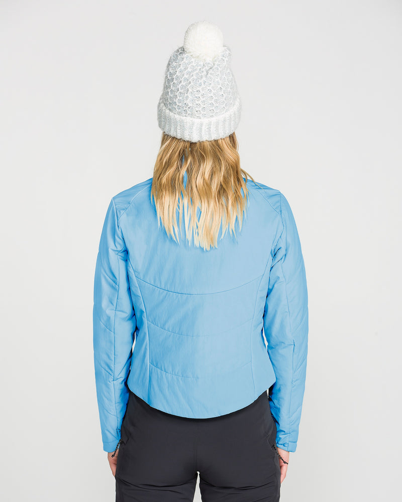 A person stands with their back to the camera, wearing the Volcom Women’s Evergreen Full Zip Jacket in light blue—featuring recycled insulation—paired with black pants and a white knit beanie with a pom-pom.