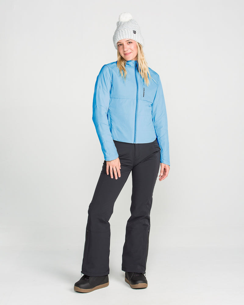A woman wearing the Volcom Women’s Evergreen Full Zip Jacket in light blue, paired with black pants, black boots, and a white knit beanie with a pom-pom, stands smiling against a plain white background.