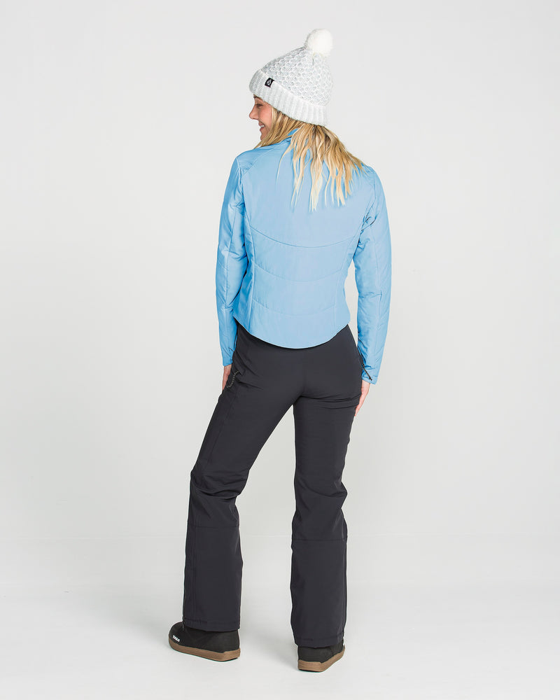 A person stands facing away, wearing a white knit beanie, black pants, and brown shoes, showcasing the Volcom Women’s Evergreen Full Zip Jacket in light blue with recycled insulation against a plain white background.