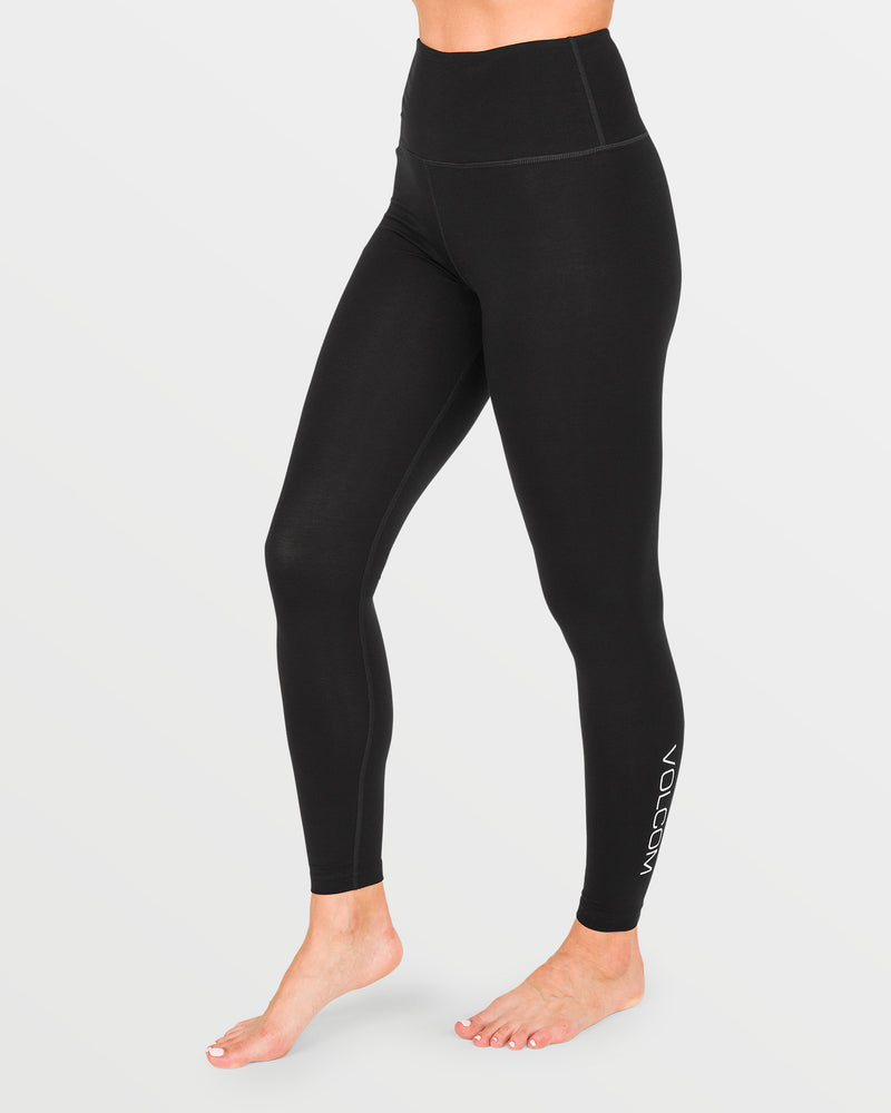 A person wearing Volcom Women’s W Merino Blend Pants stands barefoot against a white background. The high-waisted black leggings, designed for quick-dry comfort, feature “VOLCOM” written near the ankle on the left leg.