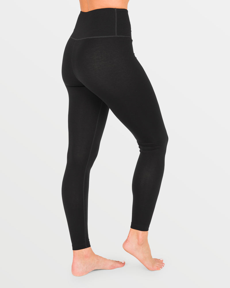 A person stands barefoot on a white background, shown from the side and back, wearing high-waisted black Volcom Women’s W Merino Blend Pants. The sleek design delivers quick-dry comfort, making these pants ideal for thermal defense layering in cooler conditions.