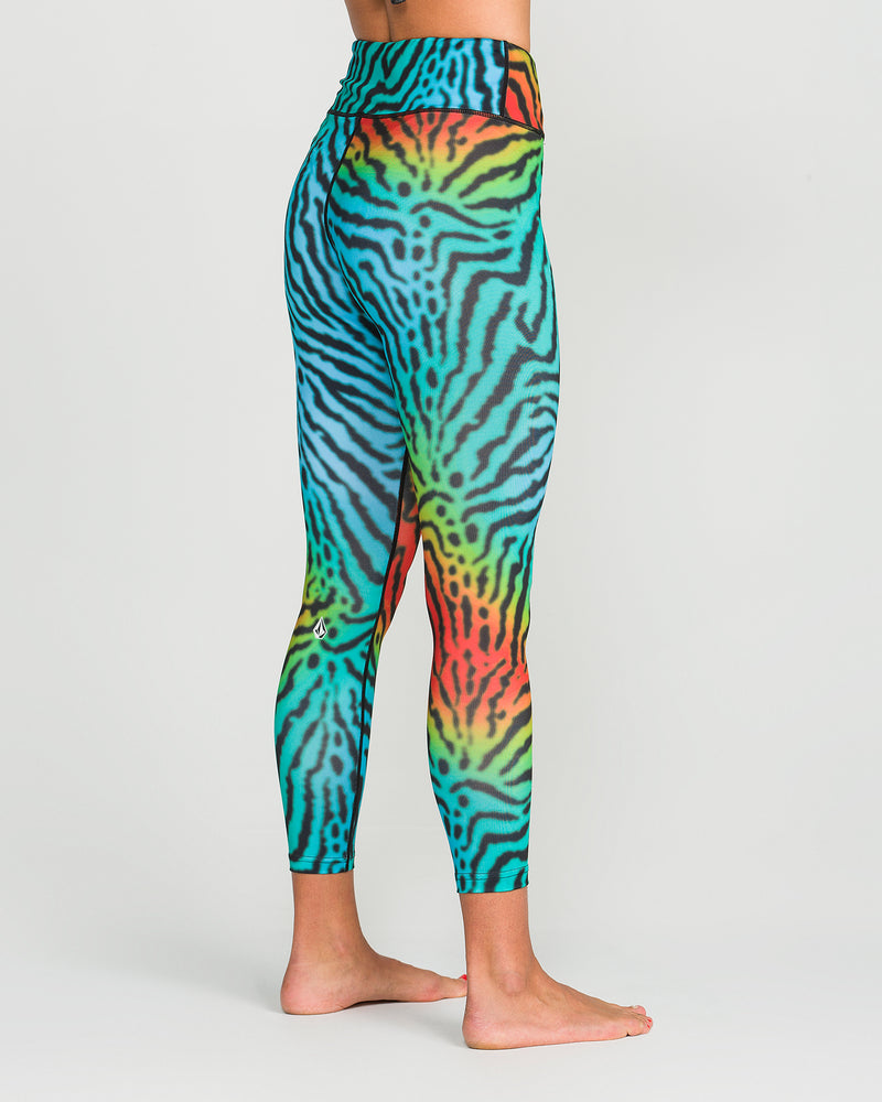 A woman stands barefoot on a plain white background, shown from the side, wearing Volcom Women’s W V-Science Pants—colorful, high-waisted leggings crafted from quick-dry knit fabric featuring a vibrant zebra-like rainbow pattern.