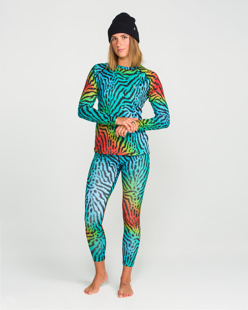 A woman stands barefoot against a plain white background, wearing Volcom’s Women’s W V-Science Pants paired with a matching long-sleeve top. The set features a vibrant wavy animal-print pattern in blue, green, red, and yellow. Crafted from quick-dry knit fabric for lasting comfort and effortless style, she finishes the look with a black beanie.