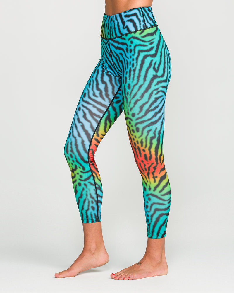 A woman stands barefoot against a plain white background, wearing Volcom Women’s W V-Science Pants—high-waisted leggings featuring a vibrant zebra-print in blue, green, yellow, and orange tones, crafted from quick-dry knit fabric.