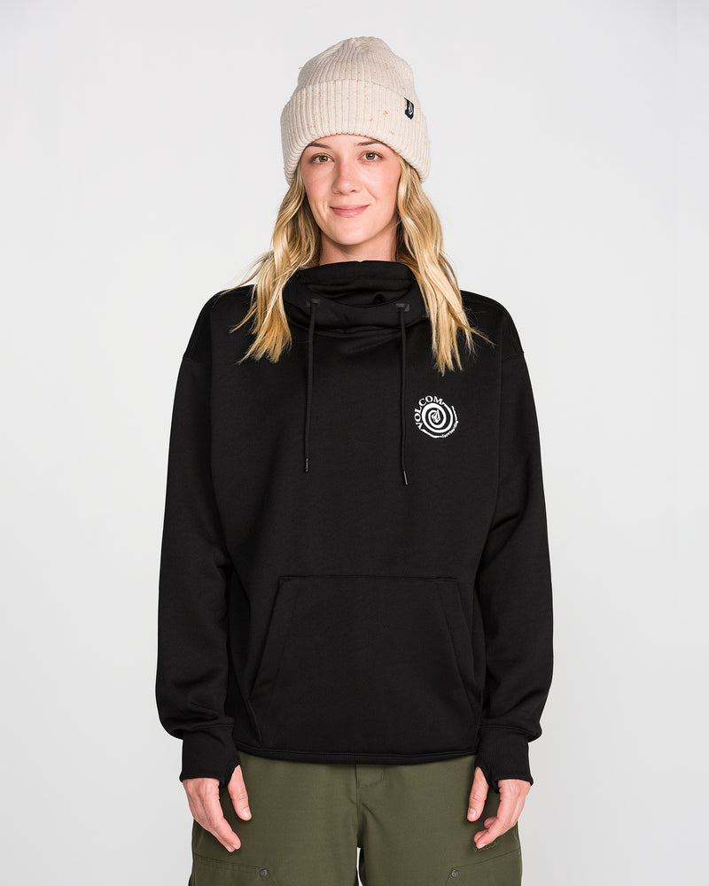 A person with long blonde hair wears a beige knit beanie, green pants, and the Volcom Women’s Riding Hydro Pullover Hoodie—a black hoodie featuring a white logo on the chest—standing and smiling against a plain white background.