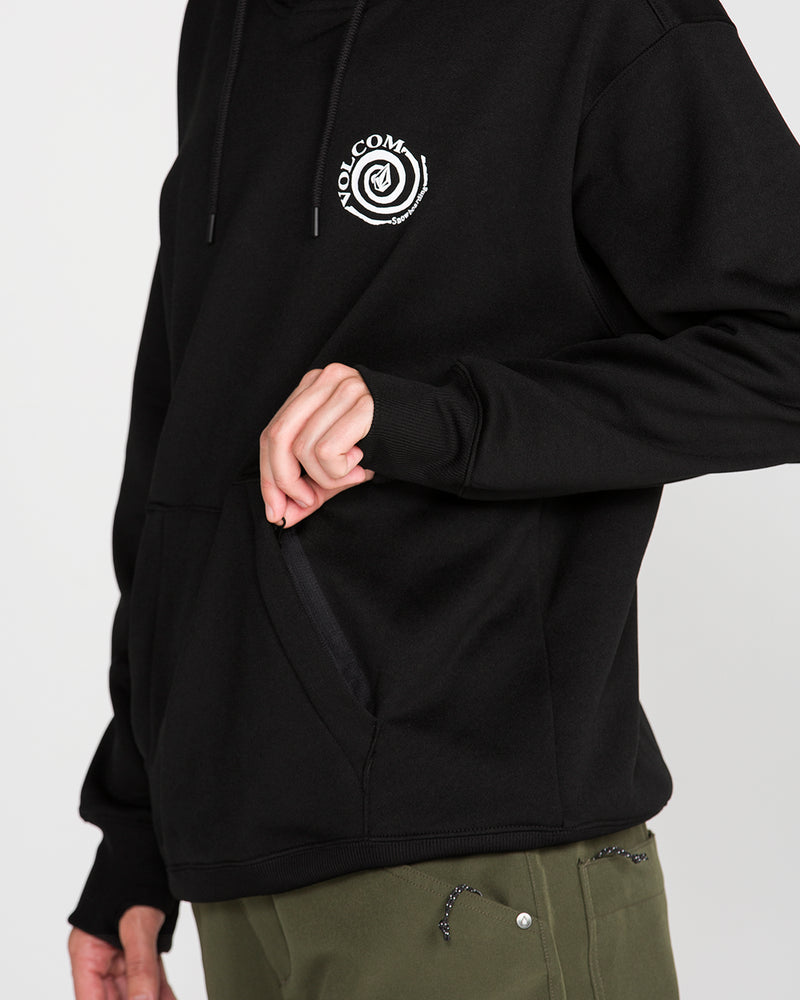 A person wearing the Volcom Women’s Riding Hydro Pullover Hoodie in black, featuring a white spiral logo, holds open the pocket with one hand. The hoodie is paired with olive green pants.