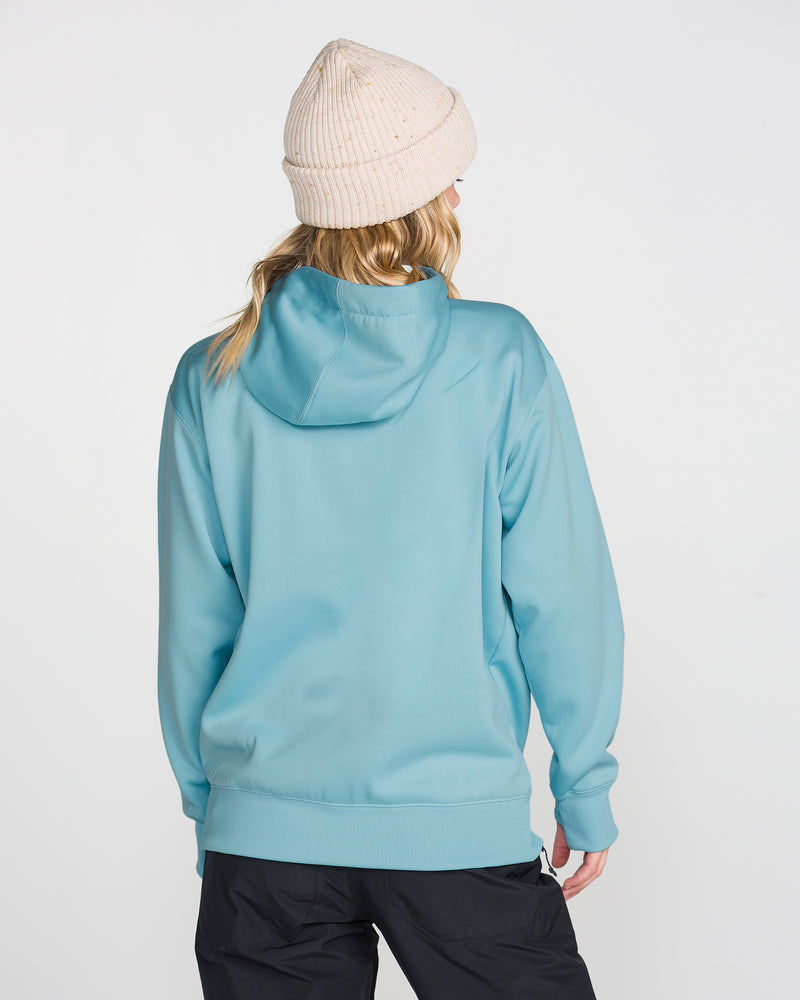 A person wearing the Volcom Women’s Riding Hydro Pullover in light blue, paired with black pants and a cream knit beanie, stands facing away from the camera against a plain white background.