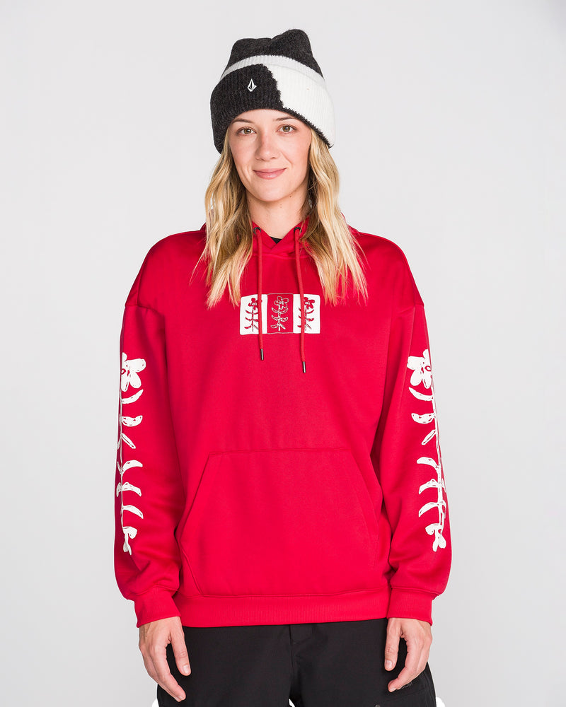 A person wearing the Volcom Women’s Essential Hydro Hoodie in red, featuring white designs on the sleeves and chest, along with black pants and a black-and-white beanie, stands against a plain light background, smiling at the camera.