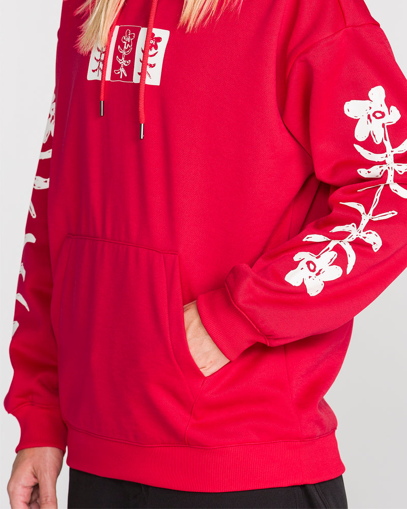 A person is wearing the Volcom Women’s Essential Hydro Hoodie in bright red, featuring white floral designs on the sleeves and chest, with their left hand in the front pocket; only the torso and part of the arm are visible.