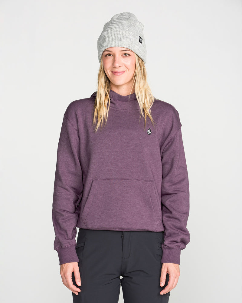 A person with long blonde hair wears a gray beanie, black pants, and the Volcom Women’s Alpine All Day Pullover—a purple cropped fleece sweatshirt featuring a cozy handwarmer pocket—while standing against a plain light background and smiling slightly at the camera.