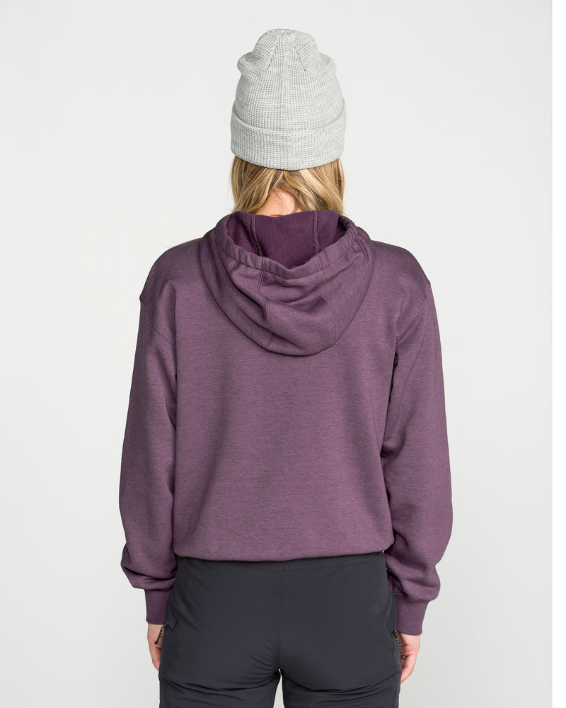 A person is shown from behind against a plain light background, wearing a gray knit beanie, black pants, and the Volcom Women’s Alpine All Day Pullover—a purple hooded sweatshirt featuring a funnel hood.